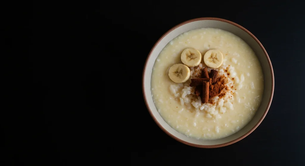Creamy arroz con leche topped with banana slices, cinnamon powder, and cinnamon sticks in a bowl on a dark background