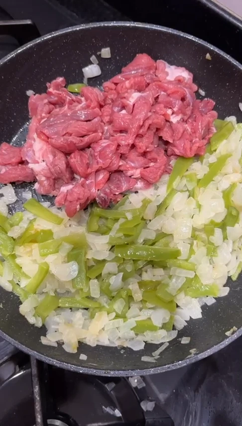 overhead view of a skillet with thinly sliced raw ribeye on one side and sautéed onions with green bell peppers on the other, starting a philly cheesesteak