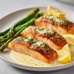 Close-up of three fillets of crispy skin salmon with lemon butter sauce, roasted asparagus, and lemon wedges on a white plate.
